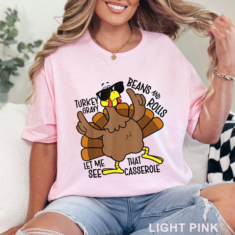 Puede incluir: Camiseta rosa claro con un dibujo de un pavo de dibujos animados con gafas de sol y gesticulando con sus alas. El texto de la camiseta dice "Turkey Gravy", "Beans and Rolls", "Let Me See That Casserole". Las palabras "Light Pink" est&aacute;n en la parte inferior.