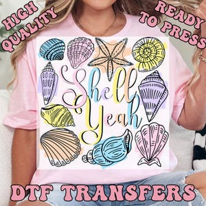 May include: A light pink t-shirt featuring a colorful graphic of various seashells and a starfish, with the words "Shell Yeah" in a playful font. The design includes blue, pink, yellow, and purple accents. The words "High Quality" and "Ready to Press" are also visible.