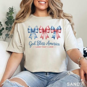 May include: Sand-colored t-shirt featuring three patriotic bow designs in red, white, and blue. The text "God Bless America - Land That I Love" is printed below the bows. The shirt is a casual style.