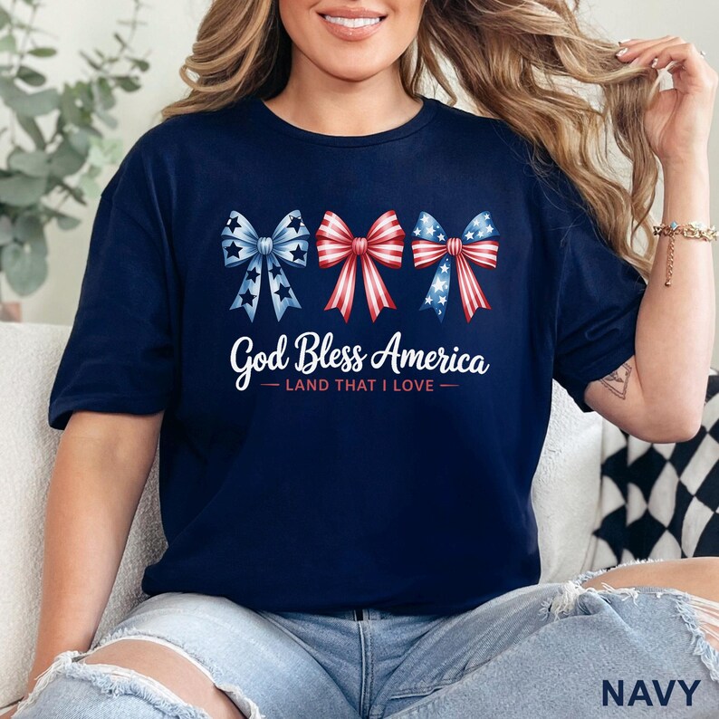 May include: Navy blue t-shirt featuring three patriotic bow designs in red, white, and blue. The text "God Bless America - Land That I Love" is printed below the bows. The shirt is a comfortable, casual style.