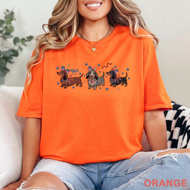 May include: Orange t-shirt featuring three dachshunds with patriotic bow ties and star accents. The dogs are brown, gray, and black, with red, white, and blue details. The word "ORANGE" is printed at the bottom of the shirt.