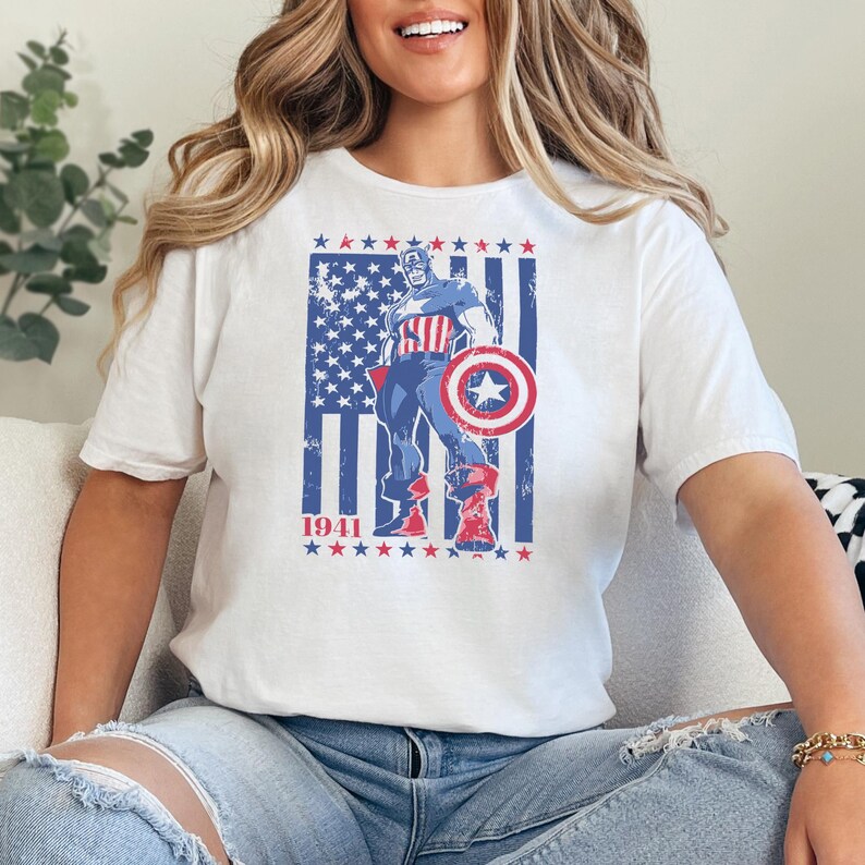 May include: White t-shirt featuring a graphic of Captain America with a shield, standing in front of a stylized American flag. The year "1941" is printed below the flag. The design uses red, white, and blue colors.