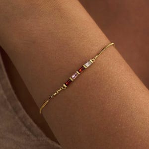 May include: A delicate gold chain bracelet featuring three small, rectangular baguette-cut gemstones in a row. The stones are coloured red, pink, and clear white, set horizontally within the thin, polished gold metal chain links.