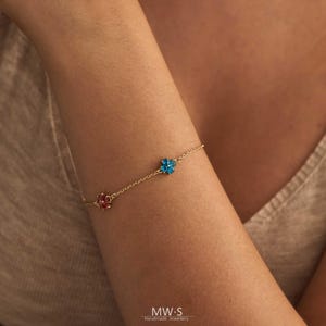 May include: A delicate gold bracelet featuring three flower charms. One charm is red, and the other is blue. The bracelet is worn on an arm, and the background is a neutral color. The bracelet is a piece of jewelry.