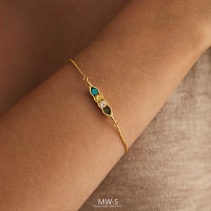 Peut inclure: Bracelet doré avec un design de gousse de pois et des pierres précieuses colorées. Le bracelet a une chaîne délicate et un charm en forme de gousse de pois avec des pierres bleues, jaunes et vertes. Bijou fait main.