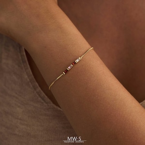 Pode incluir: Uma delicada pulseira de ouro com uma série de pedras preciosas retangulares coloridas. A pulseira inclui pedras vermelhas, rosa e brancas, presas a uma fina corrente de ouro. A pulseira é mostrada em um braço. A marca "MW-S Handmade Jewellery" é visível.