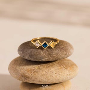 May include: A gold ring featuring three square gemstones: a clear, a dark blue, and a yellow. The ring is presented on a stack of three smooth, rounded stones. The ring is handmade jewellery.