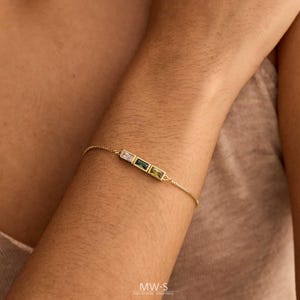 May include: A delicate gold bracelet with three rectangular gemstones in white, green, and yellow. The bracelet is worn on an arm, showcasing the jewelry's design. The bracelet is a handmade piece.