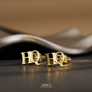 Op de afbeelding: Een paar goudkleurige metalen manchetknopen met de letters HQ in een vetgedrukt schreeflettertype. Elke manchetknop heeft een gepolijst, reflecterend oppervlak en een standaard tuimelmechanisme aan de achterkant om door knoopsgaten te bevestigen.