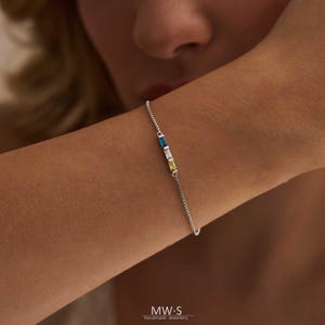 Peut inclure: Un bracelet en argent délicat orné de trois pierres rectangulaires bleues, blanches et jaunes. Le bracelet est porté au poignet et est un bijou fin. La marque "MW-S Handmade Jewellery" est visible en bas.
