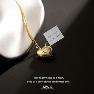 May include: A gold heart-shaped pendant on a thin gold chain. The pendant is engraved with the handwritten text "Timeless love E t M Forever," which matches the text on a small white note shown next to the heart. The text "Your handwriting, on a heart. Send us a photo of your handwritten note. MW·S Handmade Jewellery" is printed at the bottom.