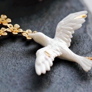May include: A white ceramic dove figurine with gold accents on the wings and tail. The dove is holding a gold floral branch in its beak. The figurine is set against a dark gray, textured surface.