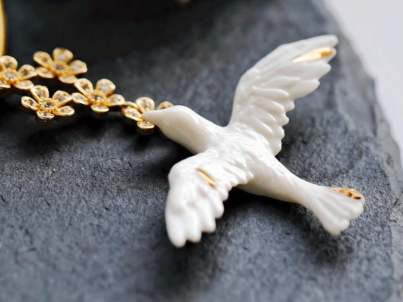 May include: A white ceramic dove figurine with gold accents on the wings and tail. The dove is holding a gold floral branch in its beak. The figurine is set against a dark gray, textured surface.