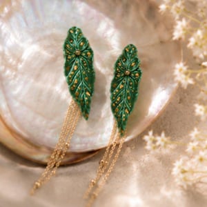 Può includere: Un paio di orecchini pendenti con ciondoli a forma di foglia verde con macchie dorate e venature in rilievo. Ogni foglia è fissata a tre delicate catenelle in metallo color oro che pendono verso il basso, creando un aspetto stratificato.