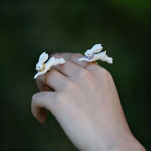 Può includere: Due anelli bianchi a forma di coniglio con fasce dorate e collari decorativi. Un anello ha un collare dorato e l'altro un collare viola. Gli anelli sono su una mano con uno sfondo verde sfocato.