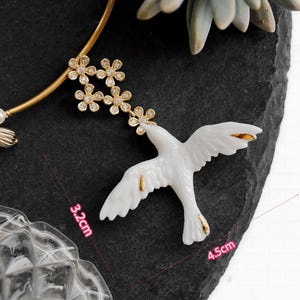 May include: A white ceramic dove pendant, 4.5 inches long, with gold accents on the wings and tail. The dove is connected to a gold-tone necklace with a floral design and a pearl accent. The pendant is displayed on a dark gray surface.