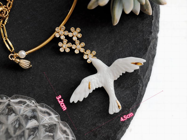 May include: A white ceramic dove pendant, 4.5 inches long, with gold accents on the wings and tail. The dove is connected to a gold-tone necklace with a floral design and a pearl accent. The pendant is displayed on a dark gray surface.