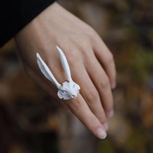 Può includere: Un anello bianco a forma di coniglio con lunghe orecchie e un viso dettagliato. L'anello ha dettagli dorati intorno agli occhi e al naso. L'anello è indossato su un dito.