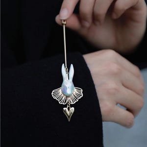 Peut inclure: Épingle dorée représentant un lapin blanc aux reflets irisés. Le lapin porte un tutu doré et un petit pendentif en forme de cœur doré. L'épingle est tenue par une personne.