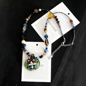 Può includere: Una collana di perline colorate con un ciondolo a forma di gatto dei cartoni animati in una mela verde. La collana include perline di varie forme e colori, tra cui blu, marrone e giallo. La collana è su una carta bianca con la scritta "NEW VOGUE".