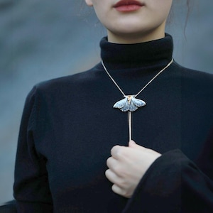 Op de afbeelding: Zilverkleurige ketting met een hanger in de vorm van een mot. De hanger heeft een gedetailleerd motontwerp met blauwe en zilveren vleugels en een klein oranje accent. De ketting wordt gedragen over een zwarte coltrui.