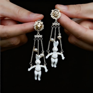 Peut inclure: Boucles d'oreilles dorées avec des clous en forme de tête de lion. Chaque boucle d'oreille comporte une chaîne pendante avec une figurine de lapin blanc et de petites perles. Les boucles d'oreilles sont présentées sur un fond sombre.
