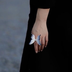 Op de afbeelding: Een witte en blauwe vlinderring. De ring heeft een vlinderontwerp met witte vleugels en blauwe accenten. De ring wordt aan een vinger gedragen, met de vlindervleugels open. De achtergrond is een wazig grijs en zwart.