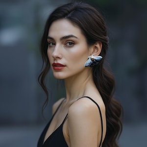 Può includere: Una donna con i capelli scuri e il rossetto rosso indossa un abito nero e orecchini a farfalla. Gli orecchini sono bianchi, neri e blu. Lo sfondo è sfocato.