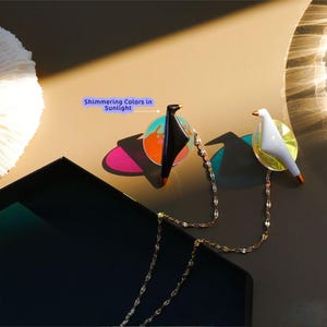 Può includere: Due spille a forma di uccello, una nera e una bianca, collegate da una delicata catena d'oro. Ogni uccello è posato su un disco colorato e iridescente. Le spille hanno becchi e zampe dorate. Il testo "Shimmering Colors in Sunlight" è visibile.
