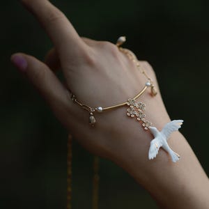 May include: A delicate gold-toned hand chain featuring a white dove pendant with outstretched wings. The chain includes small floral accents and pearl embellishments. The jewelry is designed to be worn on the hand, with a chain that wraps around the finger.
