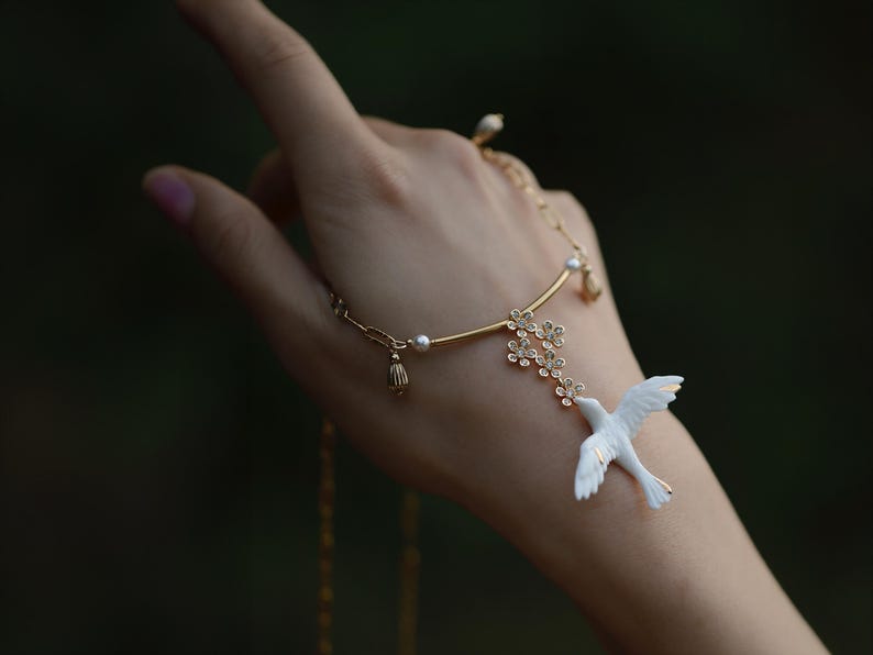 May include: A delicate gold-toned hand chain featuring a white dove pendant with outstretched wings. The chain includes small floral accents and pearl embellishments. The jewelry is designed to be worn on the hand, with a chain that wraps around the finger.