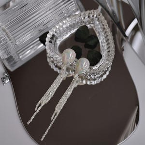Può includere: Eleganti orecchini pendenti con top iridescenti a forma di perla e fili scintillanti simili a cristalli. Gli orecchini poggiano su una superficie riflettente argentata, vicino a un piatto di cristallo a forma di cuore.