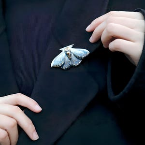 Può includere: Una spilla a forma di falena argentata e blu con dettagli dorati è appuntata su una giacca nera. La spilla presenta ali dettagliate e un design delicato. La giacca è di colore scuro e la spilla è il punto focale.
