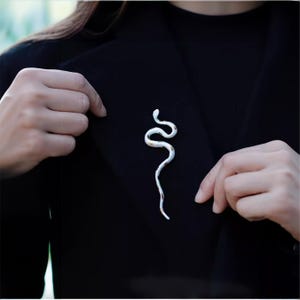 Può includere: Spilla a forma di serpente bianca e oro appuntata su una giacca nera. La spilla è una rappresentazione stilizzata di un serpente, con un corpo curvo e segni dettagliati. Un accessorio unico e accattivante.