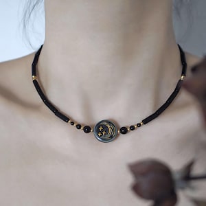 Può includere: Una collana girocollo nera con un design celeste. La collana presenta un pendente centrale circolare con una luna crescente e tre stelle dorate. La collana è realizzata con perline nere e dettagli dorati.