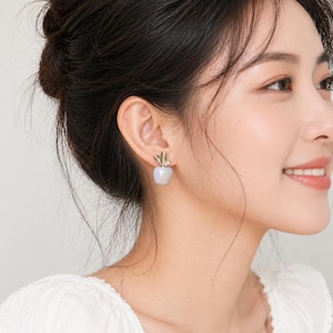 Può includere: Primo piano di una persona che indossa orecchini a forma di mela. Gli orecchini presentano un gambo e un design a foglia color oro sopra un corpo a forma di mela bianco-azzurro chiaro perlato. Un accessorio unico ed elegante.