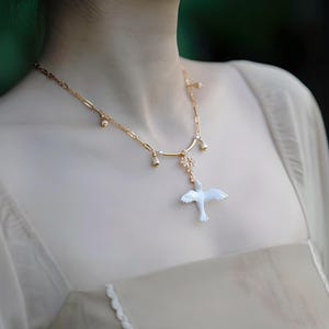 May include: A delicate gold-toned necklace featuring a pendant shaped like a bird in flight. The necklace has small, dangling accents and is worn against a cream-colored garment. The bird pendant is white with subtle iridescent details.