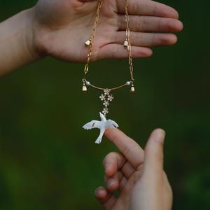 May include: A delicate gold necklace featuring a white bird pendant. The necklace has a floral design with small pearls and gold accents. The bird is perched on a finger, set against a blurred green background.