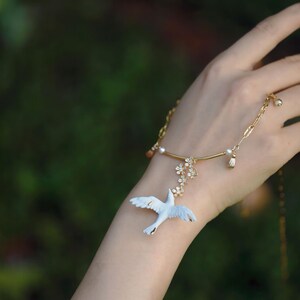 May include: A delicate gold-toned bracelet featuring a white bird pendant with outstretched wings. The bracelet includes floral and pearl accents. The background is a soft, blurred green.