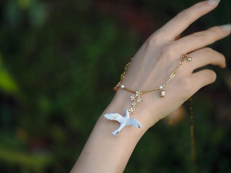 May include: A delicate gold-toned bracelet featuring a white bird pendant with outstretched wings. The bracelet includes floral and pearl accents. The background is a soft, blurred green.