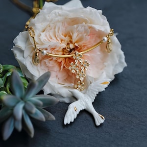 May include: A gold-toned necklace with a floral pendant and pearl accents rests on a large, light pink and white flower. A white bird figurine with gold detailing is positioned below the necklace. Green succulent plants are in the background.