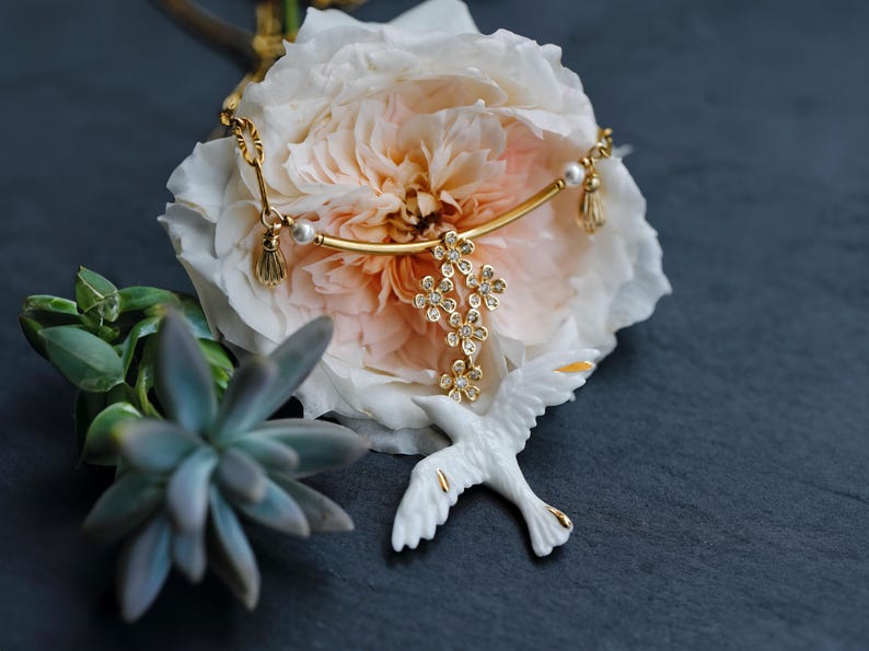 May include: A gold-toned necklace with a floral pendant and pearl accents rests on a large, light pink and white flower. A white bird figurine with gold detailing is positioned below the necklace. Green succulent plants are in the background.