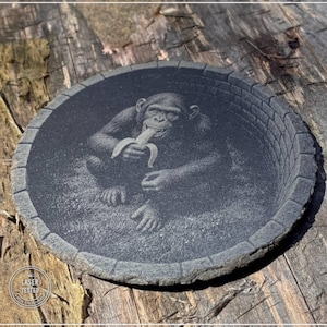 Könnte beinhalten: Ein runder Untersetzer aus dunkelgrauem Stein mit einer lasergravierten Abbildung eines Schimpansen, der eine Banane isst. Der Untersetzer hat einen strukturierten Rand, der an eine Ziegelmauer erinnert. Ein kleiner Stempel zeigt LASER TESTED.