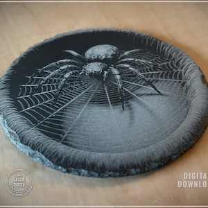 Puede incluir: Una bandeja decorativa redonda, gris oscuro, con un diseño de araña y telaraña grabado con láser. La araña está centrada en la bandeja, con su telaraña irradiando hacia afuera. La bandeja tiene una apariencia texturizada, similar a la piedra.