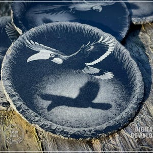 May include: Round, dark coasters with a detailed engraving of a bird in flight. The bird's wings are spread, and its shadow is visible below. The coasters have a textured edge and a natural, rustic appearance.