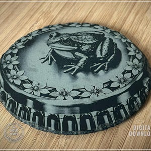 May include: A round, gray stone coaster with a detailed engraving of a frog. The coaster features a floral border and the text "LASER TESTED" and "DIGITAL DOWNLOAD".