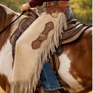 May include: Beige leather chaps featuring long fringe, intricate floral tooling on the upper yoke, and decorative silver-toned conchos along the side. The chaps are worn over blue denim jeans and brown leather boots while mounted on a horse.