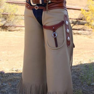 May include: Tan leather chaps with fringe along the bottom edge. The chaps have a pocket with a buckle and decorative silver studs. A brown leather belt with a large buckle is visible at the waist.
