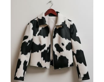 May include: A black and white cow print jacket with a collar and a zipper closure. The jacket is hanging on a wooden hanger against a white wall. The jacket's pattern resembles a cow's hide.
