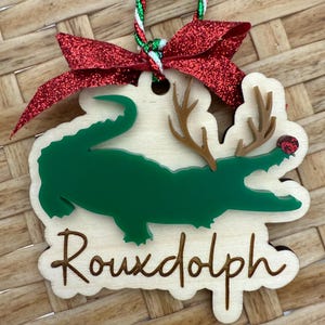 May include: Wooden Christmas ornament featuring a green alligator with antlers and a red nose, labeled "Rouxdolph." The ornament has a red glitter bow and a green, red, and white string for hanging. The background is a woven pattern.